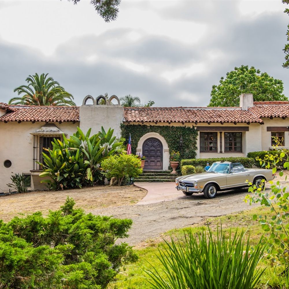 A large, white, one-story Spanish Colonial Revival house with a red tile roof, arched windows, and a classic car