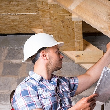 Home inspector in a white hard hat examining wooden structures, holding a clipboard and inspecting construction details.