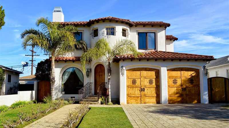 A large, two-story Mediterranean-style house with a red tile roof, palm trees, and a two-car garage.